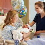 A nurse smiles at a new mother holding her newborn baby in her hospital bed