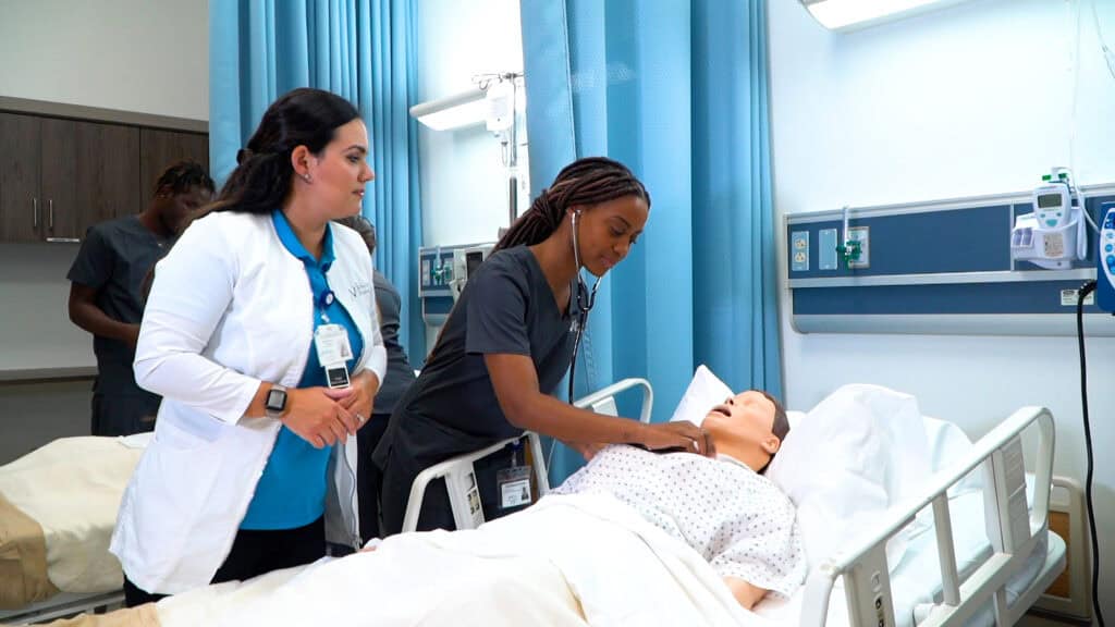 An FVI nursing instructor watches Miami and Miramar ASN nursing program students performs health checks on a patient simulator in a skills lab