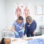 An FVI instructor guides a miramar medical assistant program student practicing hands-on skills with the assistance of another student who is laying in a hospital bed