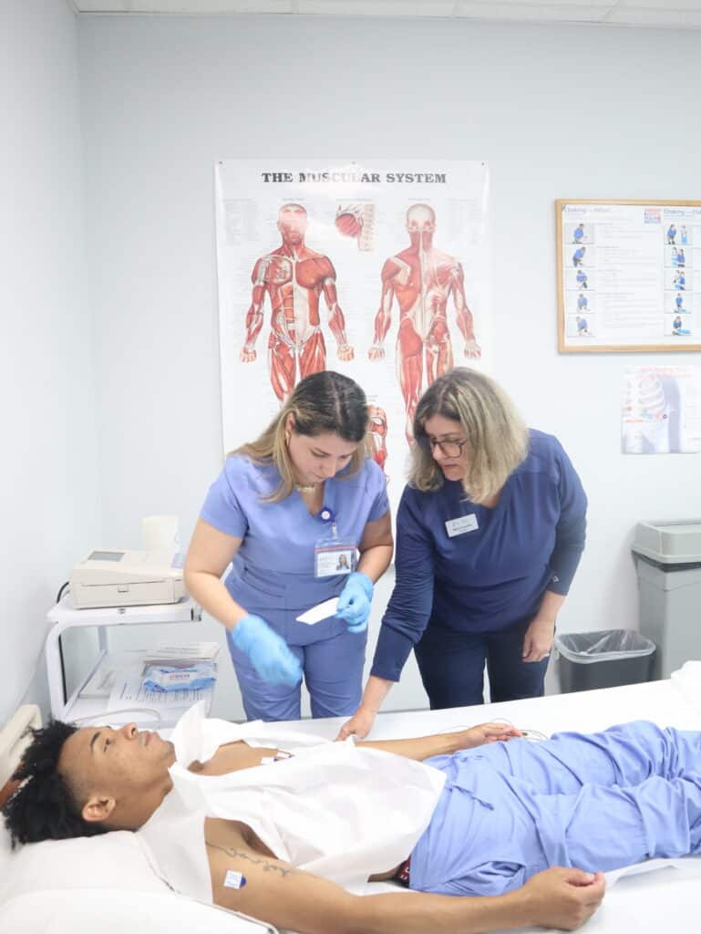 An FVI instructor guides a miramar medical assistant program student practicing hands-on skills with the assistance of another student who is laying in a hospital bed
