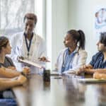 A small group florida nursing students sit around a boardroom table as they listen attentively to their teacher and lead doctor.