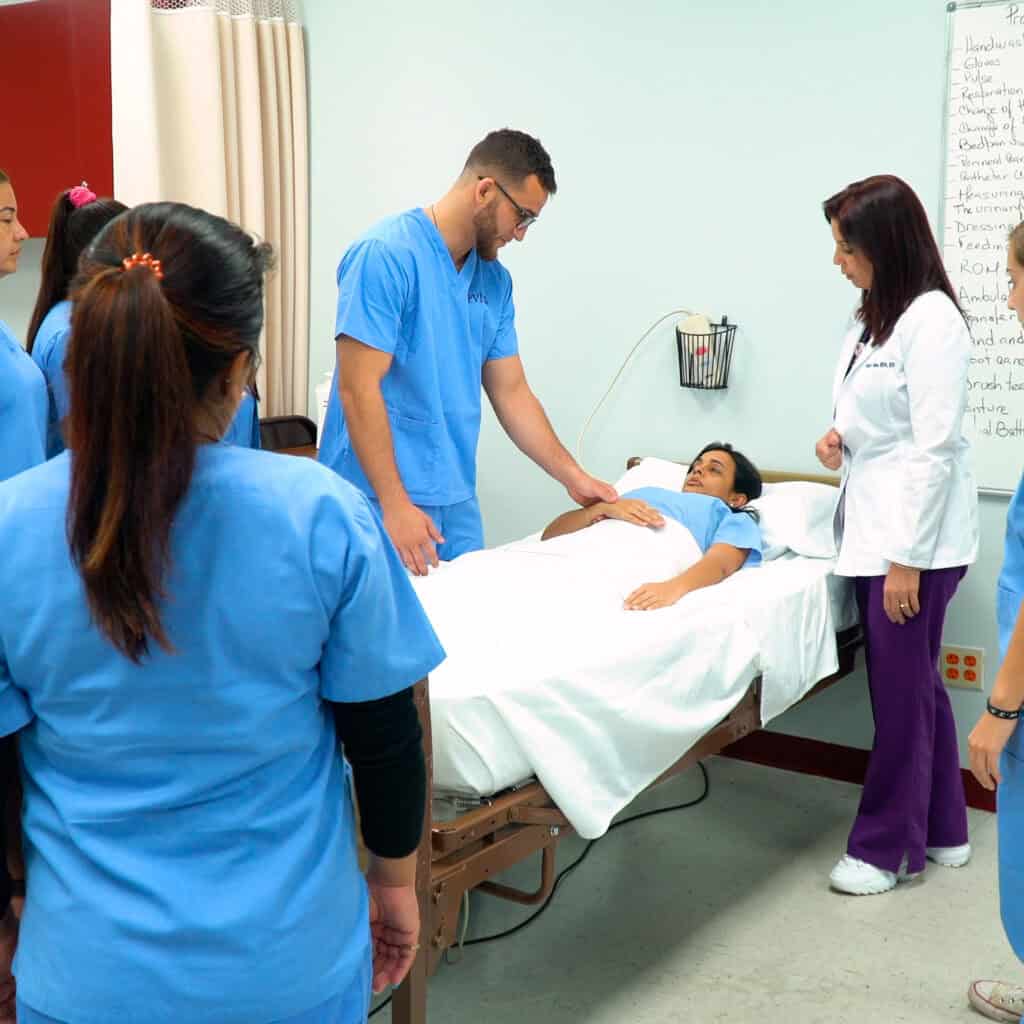 Miami patient care tech training program student simulates a real world scenario with a fellow student in a hospital bed while an instructor and fellow classmates