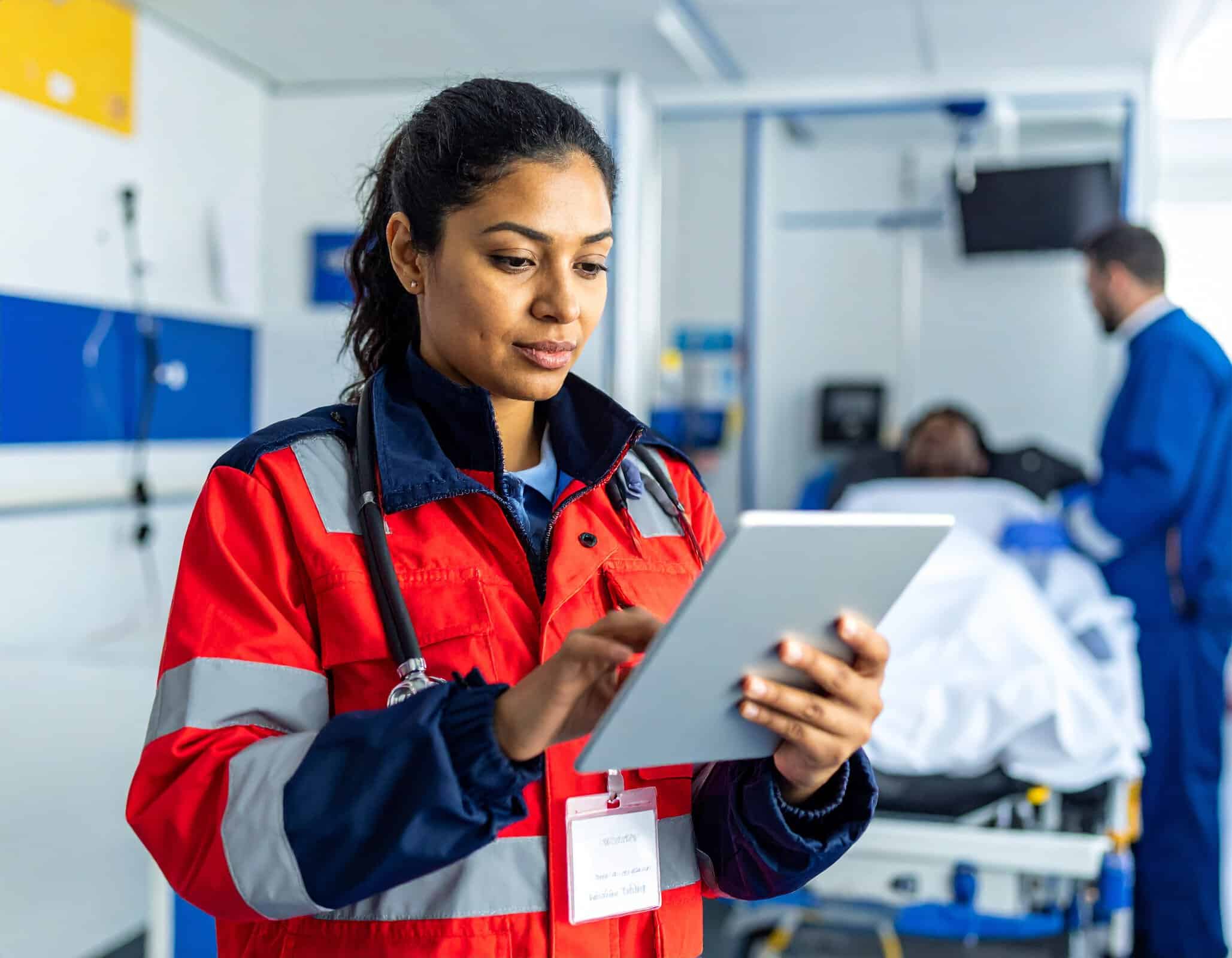 EMT professional using a tablet while monitoring a patient inside an ambulance.