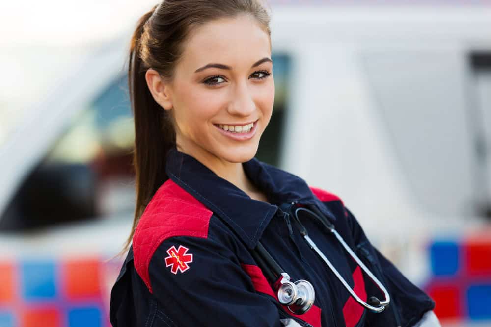 Smiling EMT professional standing in front of an ambulance with a stethoscope.