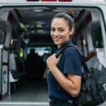 Young EMT student standing by an ambulance, smiling with a training bag on her shoulder.